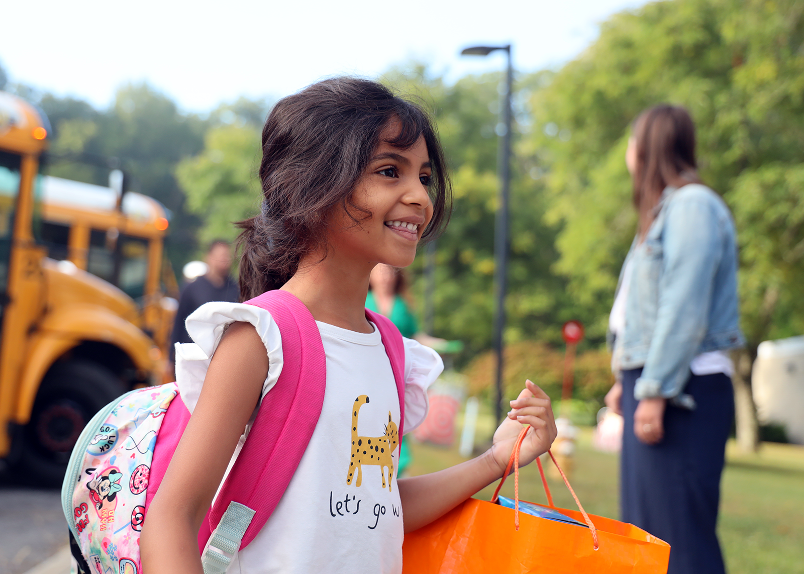 student wearing pink backpack smiling while holding orange supply bag