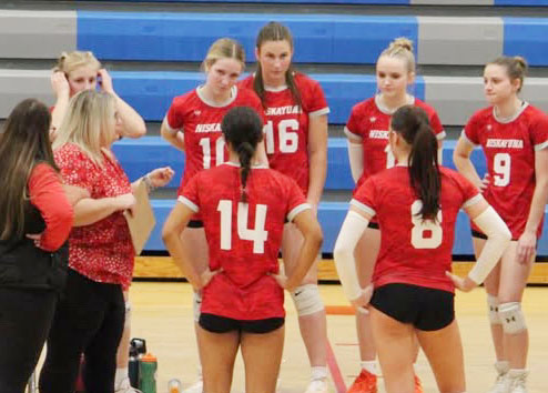 Girls Volleyball Coach Christina Coons instructs her players. Her players are wearing red uniforms with white numbers on the back and black shorts.